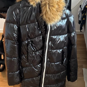 Svart pufferjacka med fuskpäls från K.Zell - Snygg svart pufferjacka från K.Zell med glansig finish och stor huva kantad med beige fuskpäls. Jackan har dragkedja framtill och är perfekt för kalla dagar. Fodret är mjukt och jackan har en oversized känsla. Riktigt statement-plagg för vintern. Den  Är helt ny och passar inte mig med lapp 