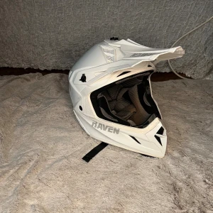 Vit motocrosshjälm från Raven - Snygg vit motocrosshjälm från Raven med coola detaljer och ventiler för extra komfort. Hjälmen har en skarp, sportig design med visir och vadderad insida. Perfekt för dig som gillar fart och äventyr. Märket syns tydligt på sidan och bak.