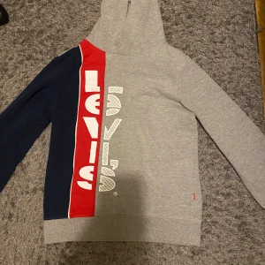 Levi's hoodie i grått, rött och blått - Snygg hoodie från Levi's med cool färgblock-design i grått, rött och mörkblått. Stor vit logga framtill, huva och långa ärmar. Perfekt för dig som gillar streetstyle och vill sticka ut lite extra.