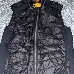 Svart dunväst från Parajumpers S - Svart dunväst från Parajumpers med quiltad framsida och baksida, dragkedja framtill och bröstficka med dragkedja. Snygga detaljer i gult och orange samt diskret logga på insidan. Perfekt för lager-på-lager under kyliga dagar.