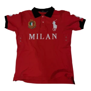 Röd pikétröja Polo Ralph Lauren Milan - Röd pikétröja från Polo Ralph Lauren med svart krage och svarta ärmslut. Stort vitt Polo-logga och MILAN-tryck framtill, broderad patch på bröstet och siffran 5 på ryggen. Klassisk piké med korta ärmar och knappar vid halsen. Perfekt för dig som gillar sportig stil.
