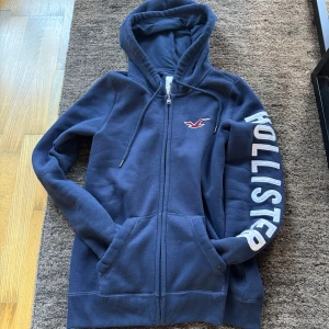 Marinblå hoodie från Hollister - Marinblå hoodie från Hollister med dragkedja, stor vit logga på ena ärmen och broderad röd fågel på bröstet. Tröjan har huva med snörning och två stora fickor framtill. Passar typ de i 175-185. Är i bra kondition förutom att texten på armen typ ser lite använd men det kan man fixa enkelt med en maskin. Priset är ej satt i sten