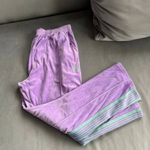 Lila mjukisbyxor med gröna ränder - Säljer ett par lila mjukisbyxor med gröna ränder längs benen och en broderad fjäril på sidan. Byxorna har elastisk midja och dragkedja vid fickan. Perfekt för chill dagar och har en riktigt skön vibe.