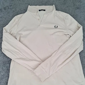 Beige långärmad pikétröja från Fred Perry - Snygg beige långärmad pikétröja från Fred Perry med klassisk krage och knäppning framtill. Tröjan har det ikoniska Fred Perry-loggan broderad på bröstet och är tillverkad i mjuk bomull. Perfekt för en clean och stilren look. Den är aldrig använd och kostar 1700kr i nypris