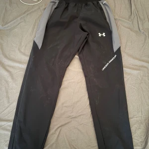 Svarta träningsbyxor Under Armour - Säljer ett par svarta träningsbyxor från Under Armour med grå paneler på sidorna. Byxorna har elastisk midja och logga på benet. Perfekta för gymmet eller löpning, riktigt sköna och snygga med sportig look.