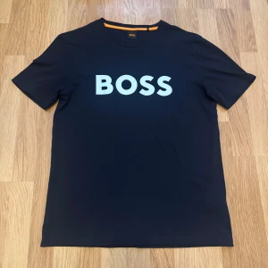 Mörkblå BOSS t-shirt med tryck - Snygg mörkblå t-shirt från BOSS med stort vitt BOSS-tryck på bröstet. Klassisk rund halsringning och korta ärmar. Perfekt för dig som gillar stilrena och trendiga plagg med tydlig logga.