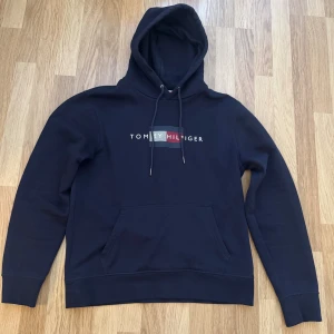 Marinblå hoodie Tommy Hilfiger - Snygg marinblå hoodie från Tommy Hilfiger med broderad logga och flagga på bröstet. Tröjan har huva med snörning, känguruficka och långa ärmar. Perfekt för dig som gillar klassisk streetstyle och vill ha något bekvämt och stilrent.
