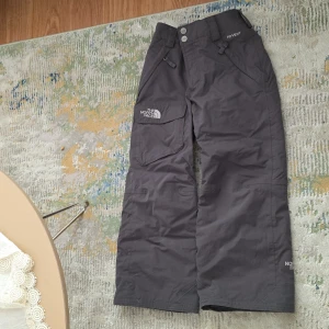 Grå vinter täckbyxor från The North Face 5år - Grå täckbyxor från The North Face med bred passform och praktiska fickor. Byxorna har justerbar midja med kardborre och tryckknappar, samt fleecefoder på insidan för extra värme. Perfekta för kalla dagar och vinteraktiviteter. För pojke barn 5 år