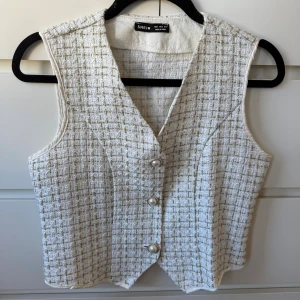 Vit/guld rutig väst med pärlknappar  - Stilren vit väst från SHEIN i rutigt tyg med inslag av guldtråd. Västen har v-ringning och stängs med tre snygga pärlknappar framtill. .