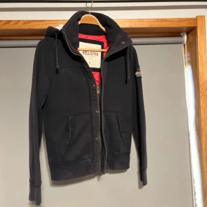 Svart teddyjacka från Hollister S - Mysig svart teddyjacka från Hollister med röd quiltad insida och vit teddyfoder i huvan. Jackan har dragkedja, stora fickor och broderad logga på ärmen. Perfekt för kalla dagar när du vill vara både varm och snygg.