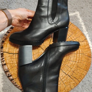 Svarta chelsea boots från Tamaris - Snygga svarta chelsea boots från Tamaris i skinn med grov sula och blockklack. Skorna har elastiska paneler på sidorna, dragkedja på insidan och rund tå. Perfekta för dig som vill ha en stilren och tidlös look med lite edge.