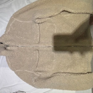 Beige teddyjacka i storlek L - Mysig beige teddyjacka med dragkedja framtill och två stora fickor. Jackan har en fluffig och varm känsla, perfekt för kyliga dagar. Passar dig som gillar en avslappnad och trendig stil.