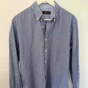 Blårutig skjorta från Morris - Snygg blårutig skjorta från Morris med klassisk button down-krage och vita knappar. Skjortan har ett broderat rosa emblem på bröstet och är tillverkad i mjuk bomull. Perfekt för dig som gillar stilrena och tidlösa plagg.