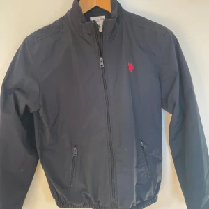 Mörkblå vindjacka från U.S. Polo Assn. - Mörkblå vindjacka från U.S. Polo Assn. med röd broderad logga på bröstet. Jackan har dragkedja framtill, två fickor med dragkedja och elastisk nederkant. Tillverkad i lätt polyester och har en klassisk, stilren look som passar perfekt till hösten!