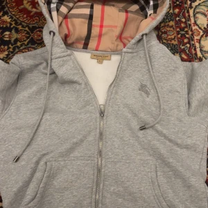 Grå hoodie från Burberry med dragkedja - Snygg grå hoodie från Burberry i storlek L med klassiskt rutigt foder i huvan. Tröjan har dragkedja framtill, snörning i huvan och två fickor. Perfekt för dig som vill ha en stilren och bekväm look med exklusiv känsla.