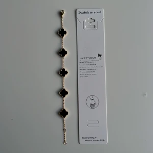 Svart och guldigt armband med klöver - Snyggt armband i rostfritt stål med fem svarta fyrklöverformade hängen, omgivna av guldfärgade detaljer. Länken är tunn och elegant, perfekt för att ge din stil en lyxig touch. Passar dig som gillar stilrena och trendiga accessoarer. Mvh clover.se