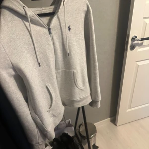 Grå hoodie från Polo Ralph Lauren - Snygg ljusgrå hoodie från Polo Ralph Lauren med dragkedja, huva och snörning. Klassisk logga broderad i marinblått på bröstet. Priset går att diskutera! 