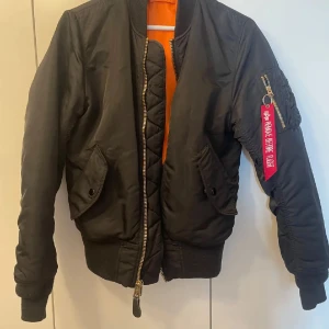 Svart bomberjacka från Alpha Industries - Säljer en svart bomberjacka från Alpha Industries med klassisk orange insida, guldfärgad dragkedja och ikonisk röd tag på ärmen. Jackan har ribbade muddar, två fickor med lock och en ärmficka med dragkedja. Perfekt för dig som gillar streetwear och snygga detaljer. Storleken är S-M