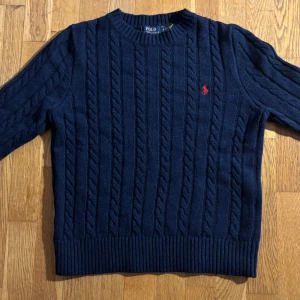Mörkblå kabelstickad tröja Polo Ralph Lauren - Kabelstickad mörkblå tröja från Polo Ralph Lauren med klassisk rund hals och röd broderad logga på bröstet. Tröjan är i ull och har ribbade muddar vid ärmslut och nederkant. Perfekt för dig som gillar preppy och stilren look.