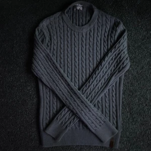 Grå stickad tröja Knowledge Cotton - Säljer en grå kabelstickad tröja från Knowledge Cotton Apparel i mjuk ekologisk bomull. Tröjan har rund hals, ribbade muddar och lång ärm. Perfekt för lager-på-lager och chill höst- eller vinterstil.