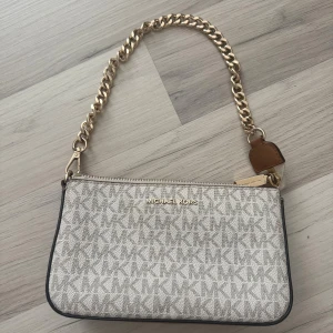  Michael Kors väska  - Snygg beige axelväska från Michael Kors med MK-logga över hela väskan. Väskan har en guldfärgad kedja som axelrem och dragkedja upptill. Insidan är fodrad med mönstrat tyg och har fack för småsaker. Perfekt storlek för det viktigaste.