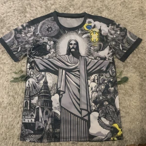 Brasil t-shirt - Unik t-shirt med stort tryck av Kristusstatyn i Rio, detaljerade svartvita mönster och inslag av gult och blått. Brasil-logga och flagga på bröstet. Rund hals och korta ärmar. Materialet är lätt och andas, perfekt för dig som gillar fotboll.