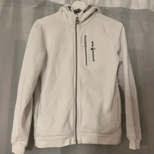 Vit hoodie från Sail Racing - Vit hoodie från Sail Racing med dragkedja framtill och bröstficka med zip. Tröjan har huva.