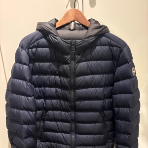 Mörkblå Colmar dunjacka med huva - Snygg mörkblå dunjacka från Colmar med huva och dragkedja framtill. Jackan har ett quiltat mönster och Colmar-logga på ärmen. Perfekt för kyliga dagar, med två sidofickor med dragkedja och en clean, sportig look.