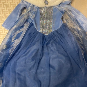 Blå Disney Frozen klänning H&M 110/116 - Säljer en magisk blå Frozen-klänning från H&M i samarbete med Disney, storlek 110/116. Klänningen har glittriga paljetter framtill, långärmade ärmar och ett fluffigt vitt fuskpäls vid halsen. Tyllkjol och transparent cape med snöflingemönster ger en riktig prinsesskänsla.