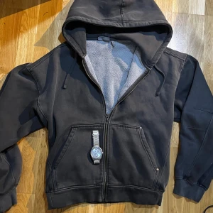 Archive hoodie - Säljer en svart hoodie med dragkedja och huva. Insidan är fodrad med mjuk fleece för extra komfort. Tröjan har stora fickor framtill och snörning i huvan. Perfekt för kyliga dagar och en avslappnad stil.