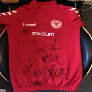 Röd Hummel half zip Kalmar FF-tröja - Röd sporttröja från Hummel med Kalmar FF-logga och flera autografer på framsidan. Tröjan har half zip, lång ärm och vita detaljer på axlarna. Perfekt för fotbollsfans som vill ha något unikt från sitt favoritlag.