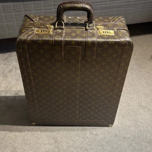 Vintage Louis Vuitton resväska monogram brun - Lyxig resväska från Louis Vuitton i klassiskt monogrammönster. Väskan är brun med guldiga detaljer och har ett hårt skal, robust handtag och dragkedja. Insidan är klädd i brunt tyg med praktiska fack och ficka med dragkedja. Skulle säga att den är mer för dekoration än att använda för att resa då den är så gammal  och tappar värde 