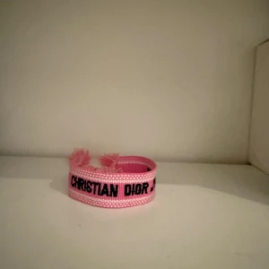Rosa armband från Christian Dior - Säljer ett snyggt rosa armband från Christian Dior med tydlig svart logotyp. Armbandet är vävt i textil och har fransiga ändar för en cool och trendig look. Perfekt accessoar för att lyfta din outfit.