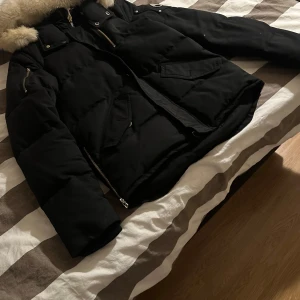 Svart pufferjacka Moose Knuckles päls - Svart pufferjacka från Moose Knuckles med fluffig naturfärgad pälskrage. Jackan har flera fickor med dragkedja och tryckknappar, samt detaljer i silver. Perfekt för kalla dagar med sin tjocka dunfyllning och lyxiga känsla. 