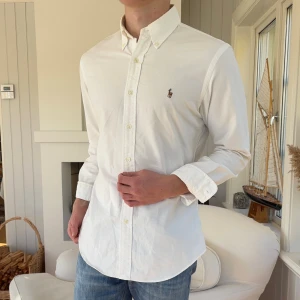 Vit skjorta från Ralph Lauren Slim Fit - Klassisk vit skjorta från Ralph Lauren i slim fit-modell. Skjortan har button down-krage, broderad logga på bröstet och är tillverkad i bomull. Perfekt passform med långa ärmar och snygga knappar framtill.