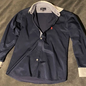 Ralph lauren skjorta 128/132 - Klassisk mörkblå skjorta från Polo Ralph Lauren med röd broderad logga på bröstet. Skjortan har långa ärmar, vita kontrastdetaljer på krage och manschetter samt knäppning framtill. Perfekt för en stilren look.