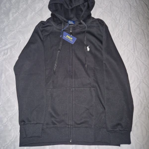 Svart hoodie från Polo Ralph Lauren - Svart hoodie från Polo Ralph Lauren med dragkedja framtill, huva med snörning och klassisk vit logga på bröstet. Mjuk bomullsblandning och två fickor framtill. Perfekt för en chill och stilren look.