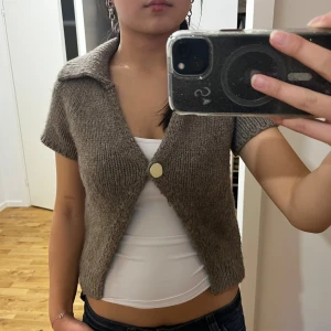 Brun stickad kofta från Zara - Kortärmad brun stickad kofta från Zara med stor krage och en snygg guldig knapp framtill. Modellen är croppad och har en relaxed fit som passar perfekt till jeans eller kjol. Supermysig och trendig för lager-på-lager-stil.