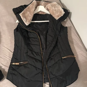 Svart dunväst med fuskpäls i huvan - Svart dunväst med dragkedja i guld och två sneda fickor med dragkedjor. Huvan är fodrad med mjuk beige fuskpäls för extra mysfaktor. Västen har en quiltad design och är perfekt för lager-på-lager under kyliga dagar. Helt oanvänd . Köpt för 1000