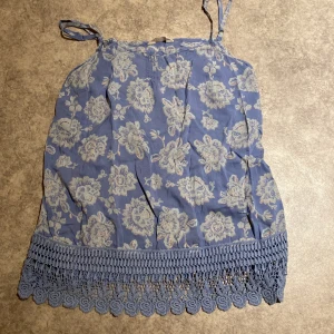 Blommig blå linnetopp från Indigo Collection - Supersöt blå linnetopp från Indigo Collection med ljusblått blommönster och knytbara axelband. Toppen har en bred virkad spetskant nertill som ger en boho-vibe. Perfekt för varma dagar och enkel att styla med jeans eller kjol.