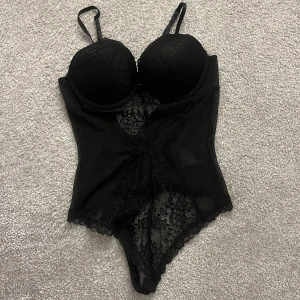 Svart spetsig body  - Säljer en svart korsett med vadderade kupor och justerbara axelband. Den har snygga spetsdetaljer framtill och meshpartier på sidorna som ger en cool look. Perfekt för att lyfta din outfit med lite edge och stil.
