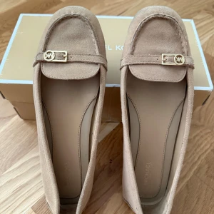 Beige mocka loafers Michael Kors - Snygga loafers från Michael Kors i beige mocka med guldfärgad MK-detalj på ovansidan. Skorna har platt sula med bruna gummipartier undertill och rund tå. Perfekt för dig som gillar klassisk och stilren design med lyxig känsla.
