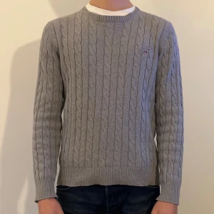 Grå kabelstickad Gant tröja   - Snygg grå kabelstickad tröja från Gant i storlek S. Tröjan har rund hals, ribbade muddar och ett litet broderat emblem på bröstet. Tillverkad i mjuk bomull, perfekt för dig som gillar klassisk stil med en modern touch. Modellen är 180 cm lång.