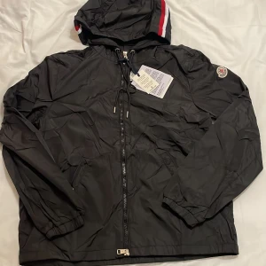 Svart vindjacka från Moncler med huva - Svart vindjacka från Moncler med huva och dragkedja framtill. Jackan har elastiska muddar, Moncler-logga på ärmen och snygga röda och vita detaljer på huvan. Tillverkad i lätt polyester, perfekt för blåsiga dagar. Snygg och sportig look.