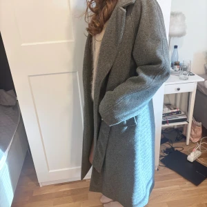 Grå lång kappa med bälte - Snygg grå kappa i klassisk modell från Gina med bälte i midjan och bred krage. Jackan är lång och har en rak passform med långa ärmar. Perfekt för dig som vill ha en stilren och tidlös look under höst och vinter.