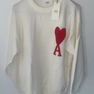 Vit stickad tröja från Ami Paris - Superfin vit stickad tröja från Ami Paris med röd hjärt- och A-detalj på bröstet. Tröjan har rund halsringning, ribbade muddar och en avslappnad passform. Perfekt för dig som vill ha en stilren men ändå unik look.