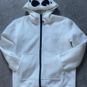 Vit gogglejacka från C.P. Company - Vit gogglejacka från C.P. Company med ikoniska svarta glasögon i huvan och broderad logga på bröstet. Jackan har dragkedja framtill, två stora fickor och är tillverkad i mjukt material. Perfekt för dig som vill sticka ut med en unik streetwear-look.tröjan är inte äkta och det är en knapp som har lossnat men det är inget som syns