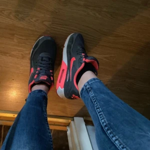 Nike Air Max 90 grå och neonröda - Nike Air Max 90 sneakers i grått med neonröda detaljer och vit sula. Klassisk siluett med synlig Air Max-dämpning, snörning och rund tå. Ovandel i textil och syntet, insida i neonrött. Perfekta för dig som vill sticka ut med färg.