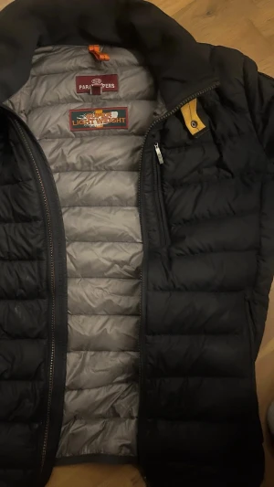 Svart pufferjacka från Parajumpers - Säljer en svart pufferjacka från Parajumpers med gul detalj vid kragen och dragkedja framtill. Jackan är superlätt och har en snygg quiltad design samt praktisk innerficka märkt 'Security Pocket'. Perfekt för kalla dagar och riktigt stilren.