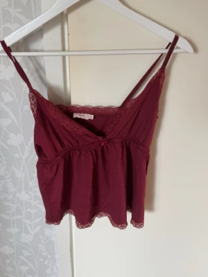 Vinröd spetsdetaljerad topp från Nelly - Säljer en vinröd topp från Nelly med tunna axelband och v-ringning. Toppen har spetsdetaljer längs urringningen och nederkanten samt en liten rosett framtill. Mjuk och lätt i materialet, perfekt för lager på lager eller som statement-plagg.Säljs pågrund av att den inte passar mig❤️
